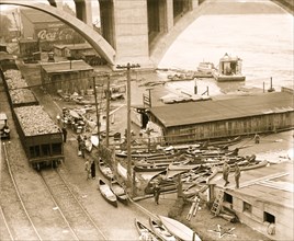 Canoes abound as flood waters lap the shore under  concrete bridge and beside railroad hopper cars. 1924