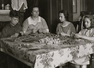 Cottage Industry shows family stringing material in piecework done at home. 1924