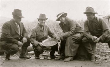 Group portrait of a woman and three men, crouched outside, smiling; one of the men is displaying a dish of panned gold 1910