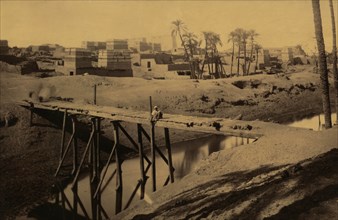 Man seated on wooden bridge over canal, ruins and buildings in background 1880