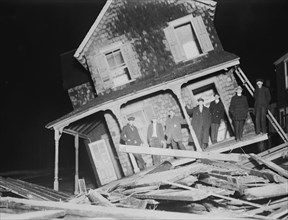 Men stand on Porch of a Sea bright Cottage torn from its foundation and tilted at a 45 degree angle.