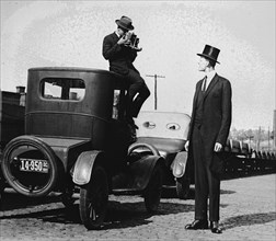 Photographer Mounts himself on the roof of  a car to shoot a pictures of an exceedingly tall men in a top hat. 1922