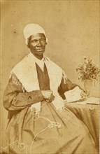 Sojourner Truth, three-quarter length portrait, seated at table with knitting and book 1864