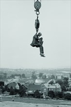 Still Photographer hangs suspended high above the ground on the hook of a large crane. 1923