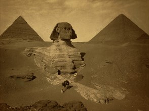 Three men, one with camel, on paw of the Sphinx, two pyramids in background. 1880