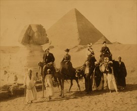 Three women and a man in western attire, seated on camels, several local men stand before the camels holding the reins, a pyramid and the sphinx in background 1880
