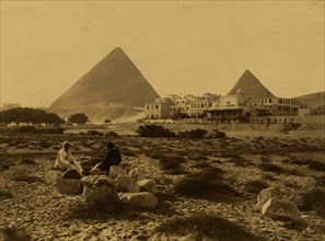 Two people sitting on rocks in an area of sparse vegetation in front of the Mena House Hotel, two pyramids in background. 1880
