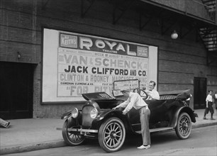 Two performers work on their car, one in the driver' seat and the other looks under the hood; Van & Schenk