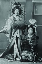 Two Young Beautiful Japanese Geishas accoutered in the ceremonial costumes and hairstyles of ancient Japan with Kimono, Combs and Obi
