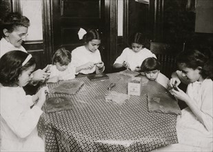 Woman and children around a table making chains for handbags 1912