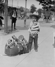 Young Cowboy, "Fog Horn Clancey" stand beside saddle bag and lasso 1923