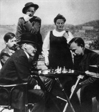 V,i, lenin (in bowler hat) plays chess with a, a, bogdanov while visiting a,m, gorky (in background, in wide-brimmed hat, standing next to natalya bogdanova), zinovll peshkov, who was standing behind ...