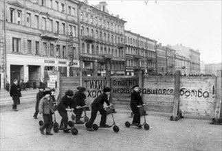 world war ll: 'drive slowly! unexploded bomb! danger!' says the notice painted on the fence, in the foreground leningrad children have organized scooter races, they have seen so much in their young li...