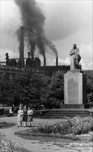 A statue of joseph stalin in the garden of victory square in the industrial city of stalinsk, the iron and steel plant is in the background, spewing smoke, early 1950s.