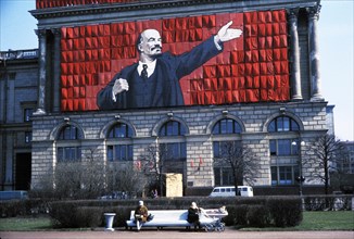 A banner of lenin on the side of a building overlooking two elderly women sitting on a park bench in leningrad, ussr, 1960s.
