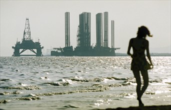 A woman in silhouette walking along shikhov beach on the shore of the caspian sea with an off-shore oil rig under construction in the background, baku, azerbaijan, october 2002.