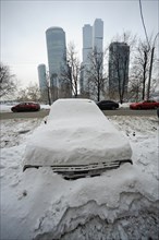 Moscow, russia, february 4, 2010, abandoned car buried in snow off a road, with the moscow international business centre (mibc) skyscrapers in the background, russia is launching a cash-for-bangers sc...