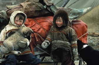 Two children of siberian reindeer herders with a puppy sitting by a packed sled, ready to start a long journey, 1990s.