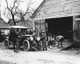 An early touring car stops at a gas station for repairs. No location, early 1900s.