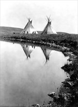 Edward S. Curits Native American Indians -  Two tepees reflected in water of pond, with four Piegan Indians seated circa 1910.