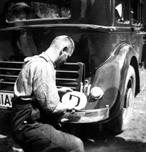 Life in Germany circa 1930s - German man affixing a country sticker to his car bumper.
