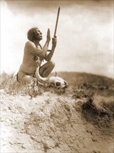 Edward S. Curits Native American Indians - Slow Bull, squatting, wearing breechcloth, holding pipe with mouthpiece pointing skyward, buffalo skull at his feet circa 1907.