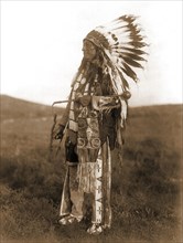 Edward S. Curtis Native American Indians - High Hawk, standing, facing left, in ceremonial dress, war bonnet and holding a coup stick circa 1907.