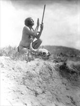 Edward S. Curits Native American Indians - Slow Bull, squatting, wearing breechcloth, holding pipe with mouthpiece pointing skyward, buffalo skull at his feet circa 1907.