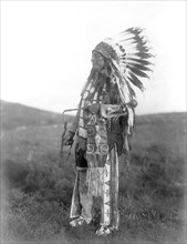 Edward S. Curtis Native American Indians - High Hawk, standing, facing left, in ceremonial dress, war bonnet and holding a coup stick circa 1907.