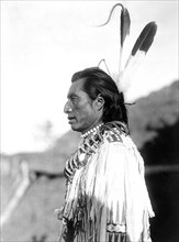 Edward S. Curtis Native American Indians - Crow's Heart wearing a buckskin shirt, two eagle feathers in his hair and necklaces, one of bear claws circa 1908.