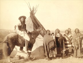 Four Lakota women standing, three holding infants in cradleboards, and a Lakota man on horseback, in front of a tipi, probably on or near Pine Ridge Reservation. 1891.