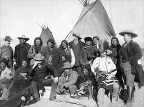Group of White Americans and Lakota (Brulé, Miniconjou, and Oglala) men standing and sitting in two rows in front of tipis..