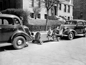 A boy occupies a parking spot in his home made go-kart circa 1937.