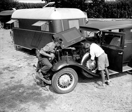 Man and his son looking under the engine and working on their car at a trailer camp circa 1937.