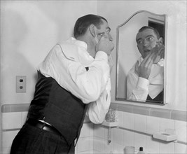 Man in modest apartment bathroom looks in the mirror as he shaves himself circa 1938.