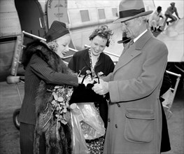 Senator William Gibbs McAdoo, of California, comes to the aid of Miss Marion Weldon, Paramount starlet, as she searches for her beauty aids before greeting the throng on her arrival at Washington Airp...