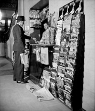 Man paying worker for a newspaper at indoor newspaper stand (news stand) circa 1940.