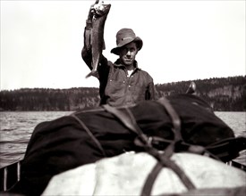 A man in a canoe, holding up a fish circa 1909.