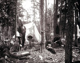 A camp site in a forest, with a tent, a man standing and another preparing a meal. Hats and boots are drying on the stumps of saplings that had been cut to support the tent circa 1909.