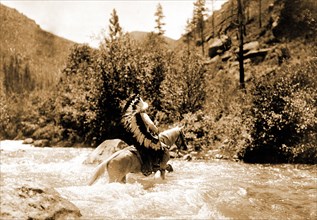 Edward S. Curtis Native American Indians - Bullchief, wearing warbonnet, crossing shallow rapids on horseback circa 1905.