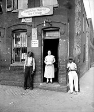 People stand outside of a small market in a slum area of Washington D.C. with Southern Dairies Ice Cream and Prince Albert ads on brick wall  circa 1935 .