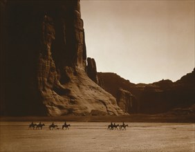 Edward S. Curits Native American Indians -  Seven Navajo riders on horseback and dog traveling against a background of canyon cliffs circa 1904.