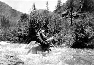 Edward S. Curtis Native American Indians - Bullchief, wearing warbonnet, crossing shallow rapids on horseback circa 1905.