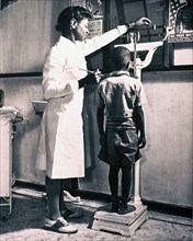 African American child is standing on a scale being weighed by a school nurse.