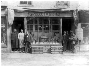 Mail carrier and six other men posed standing at entrance to shop. S.J. Gilpin shoe store