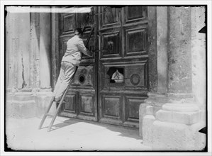 A man on a ladder is locking the locking the Church of the Holy Sepulchre, priest looking out the door ca. 1900