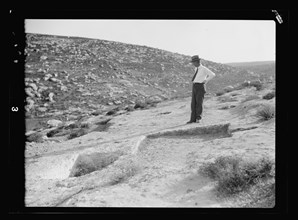 A man looks over an archelogical dig site and admires a primitive winepress showing treading-out vat ca. 1920