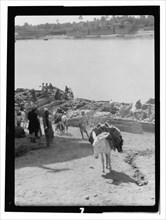 Boats being unloaded in Iraq with goods being packed on mules at a river with the city in the background ca. 1932
