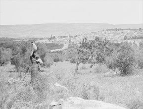 Woman holding a baby walking with jar on her head, Nazareth in the distance ca. 1925