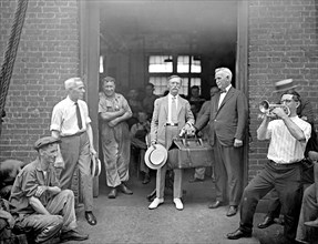 Interesting group of men, one with a suitcase, another playing a trumpet or bugle, outside a garage ca. between 1910 and 1920
